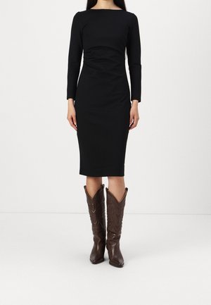 Kobieta w dopasowanej czarnej sukience midi z długim rękawem, w połączeniu ze średniej wysokości brązowymi skórzanymi kowbojskimi butami, stojąca na jednolitym białym tle.