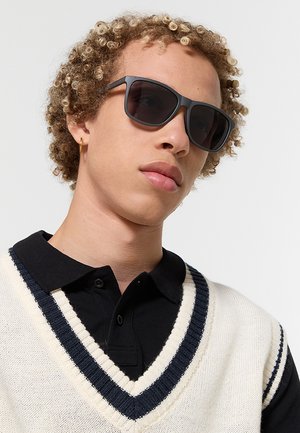 Giovane con capelli ricci, indossa occhiali da sole scuri, un piccolo orecchino a cerchio, una polo nera e un gilet bianco con rifinitura blu navy.