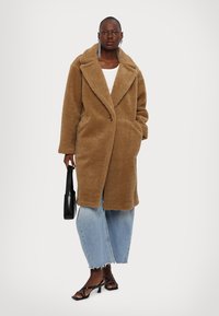 Bruine oversized teddyjas met gesneden revers, een sluiting met knopen aan de voorkant, zijzakken, gedragen over een witte top en blauwe wijde jeans.