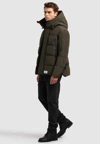 Paminkštinta alyvuogių žalia striukė su aukštu apykakliu ir gaubtu, juodais užtrauktukais ir šoniniais kišenėmis, derinama su tamsiomis džinsais ir juodais batais.