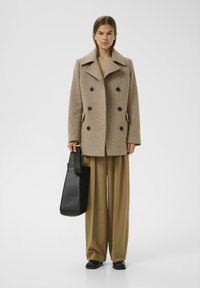 Cappotto doppiopetto in misto lana beige con bottoni neri, ampie rever e tasche laterali. Abbinato a pantaloni beige a gamba larga e una borsa nera.
