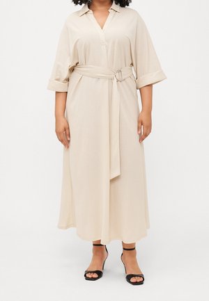 Robe chemise beige mi-longue jusqu'aux genoux, avec col, manches retroussées et ceinture assortie avec une boucle argentée. Portée avec des sandales noires à talons.