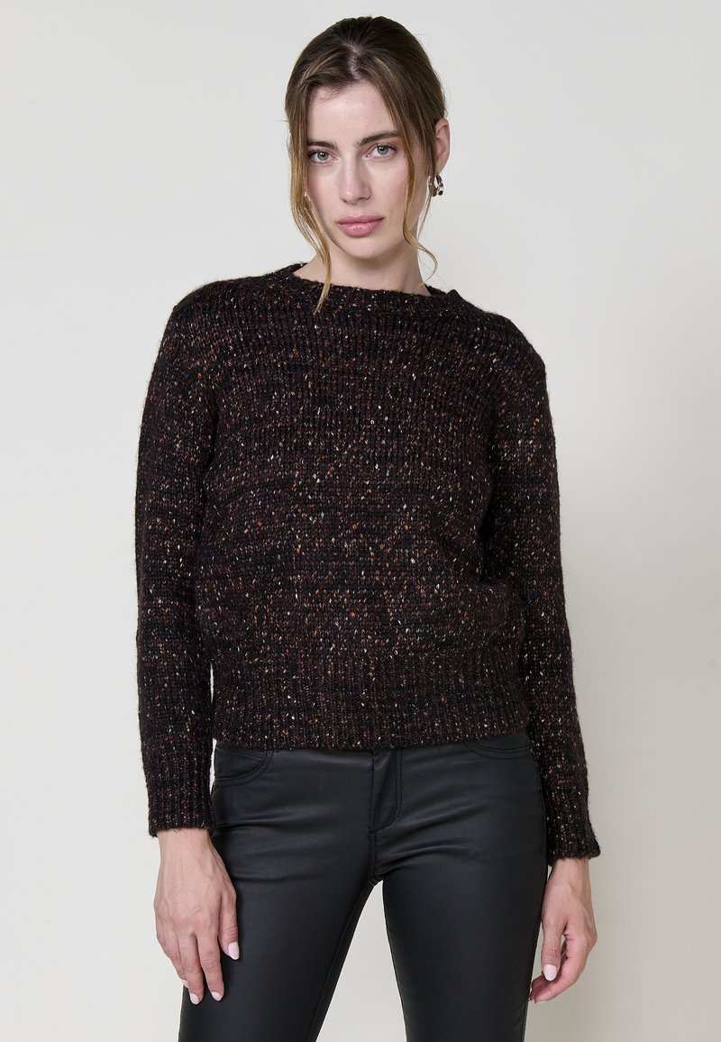 Dunkel gestrickter Pullover mit strukturiertem Muster, weitem Rundhalsausschnitt und langen Ärmeln, kombiniert mit schwarzen, figurbetonten Hosen.