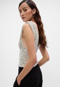 Top bez rukava s šljokicama i dubokim V-izrezom, ukrašenim dizajnom, srebrnim i iridescentnim bojama, prilagođeni kroj uparen s crnim hlačama.