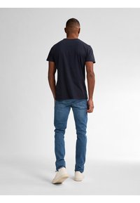 Muž stojící zády, oblečený v černém tričku, modrých džínách a bílých teniskách na neutrálním světle šedém pozadí.