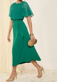 Vestido midi de gasa verde con mangas cortas y fluidas, cintura ajustada y un cinturón a juego. Accesorios con un bolso de mano texturizado dorado y sandalias de tiras.