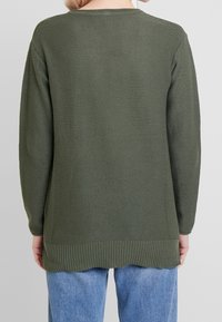 Maglione a maglia verde con texture, maniche lunghe, orlo a coste e spacco laterale. Indossato sopra jeans azzurri chiari.