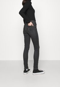 Jeans negros ajustados con una textura suave, que cuentan con un diseño estándar de cinco bolsillos y un logo sutil en el bolsillo trasero. Combinados con zapatillas negras.
