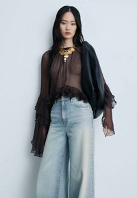 Sheer black blouse with ruffled sleeves, paired with light blue wide-leg jeans. The outfit is complemented by a chunky gold necklace and a black jacket.