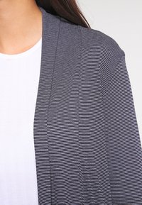 Pruhovaný cardigan v tmavě modré a bílé barvě, vyrobený z lehkého materiálu s otevřeným předním designem. Nosený přes hladký bílý top.