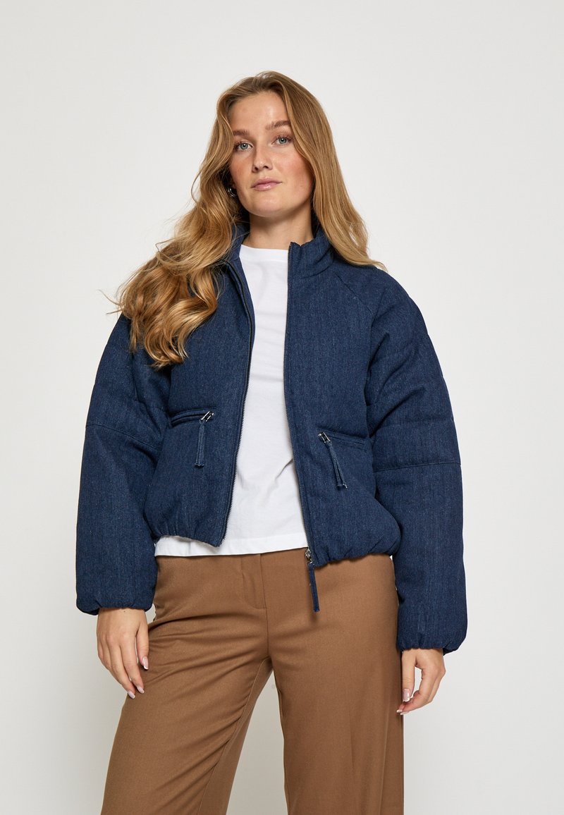 Jūras zilas pufīgā jakas ar stāvo apkakli, rāvējslēdzēja priekšpusi un divām sānu kabatām ar rāvējslēdzēju. Audumam ir teksturēta virsma.