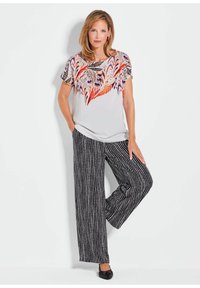 Korte mouw top met kleurrijke veerprint op een lichte achtergrond, gecombineerd met wijde, gestreepte zwart-witte broek. Zwarte schoenen.