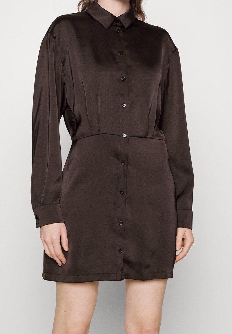 Robe chemise en satin marron avec manches longues, boutonnée devant, et taille cintrée. Dotée d’un col classique et d’un léger éclat.