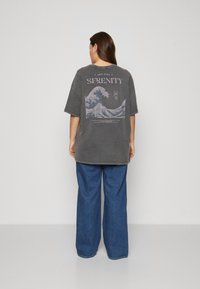 Camiseta gráfica de corte oversized en algodón gris oscuro, con un diseño de ola y el texto "SERENIDAD" en la parte posterior; combinada con jeans de corte amplio en azul.