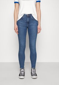 Vaqueros skinny de denim azul con cintura alta, que cuentan con cinco bolsillos y una sutil textura desgastada, combinados con zapatillas altas negras.