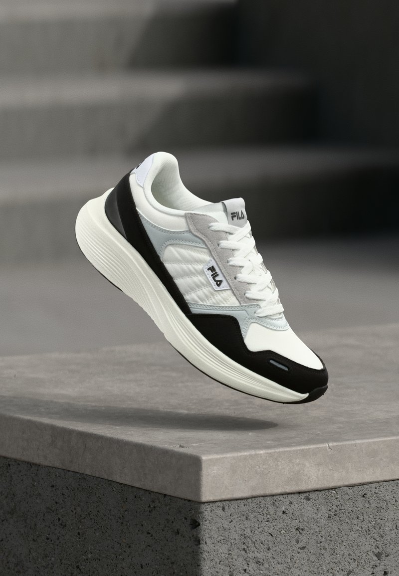 Λευκό και μαύρο αθλητικό παπούτσι FILA με κορδόνια, αιωρούμενο πάνω από μια τσιμεντένια άκρη με φόντο ένα θολό γκρι φόντο.