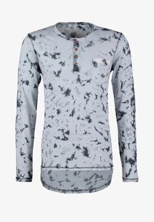 Langärmliges, hellblaues Hemd mit einem Batikmuster, geknöpftem Kragen, schwarze Jeans und graue Sneakers, mit einem grauen Rucksack über einer Schulter.