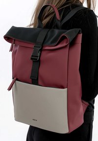 Zaino roll-top rosso e nero con una tasca frontale grigia. Realizzato in materiale elegante, dotato di spallacci regolabili e un compartimento con zip.