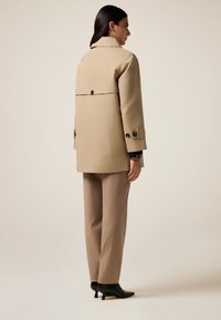 Beige waterdichte jas met een gestructureerd ontwerp, knopenaccenten op de mouwen en een detail op de rug. Gecombineerd met beige broeken en zwarte hakken.