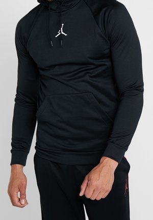 Uomo che indossa una felpa nera a maniche lunghe con cappuccio e logo Jumpman bianco, abbinata a pantaloni neri con logo Jumpman rosso sulla coscia.