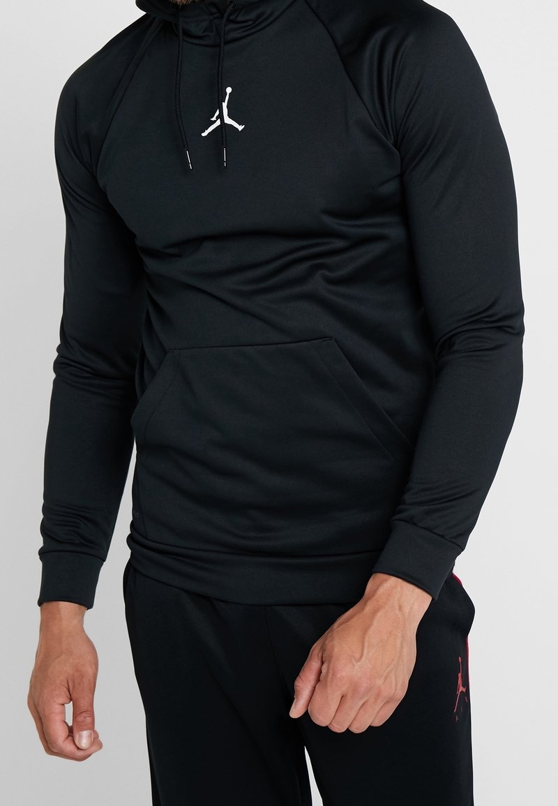 Homme portant un sweat à capuche à manches longues noir avec logo Jumpman blanc et pantalon noir assorti avec logo Jumpman rouge sur la cuisse.