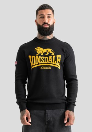 Homme avec barbe portant un pull noir Lonsdale London avec un logo lion jaune et un patch Union Jack sur la manche gauche, debout devant un fond clair.