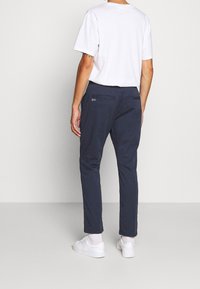 Homme portant un t-shirt blanc, un pantalon slim bleu marine avec poches arrière, des chaussettes blanches et des baskets blanches, debout devant un fond uni.