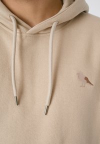 Beige Kapuzenpullover aus strukturiertem Stoff, mit einem kleinen gestickten Design eines braunen und rosa Vogels sowie silberfarbigen Metallenden an den Kordeln.