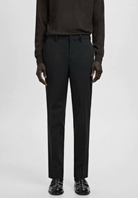 Mężczyzna ubrany w czarne spodnie garniturowe, czarne mokasyny i ciemnobrązową koszulę z długim rękawem, stojący na tle jednolitego jasnoszarego tła.