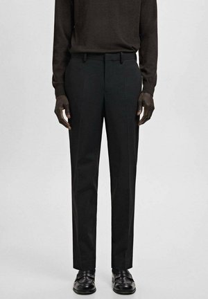 Mężczyzna ubrany w czarne spodnie garniturowe, czarne mokasyny i ciemnobrązową koszulę z długim rękawem, stojący na tle jednolitego jasnoszarego tła.