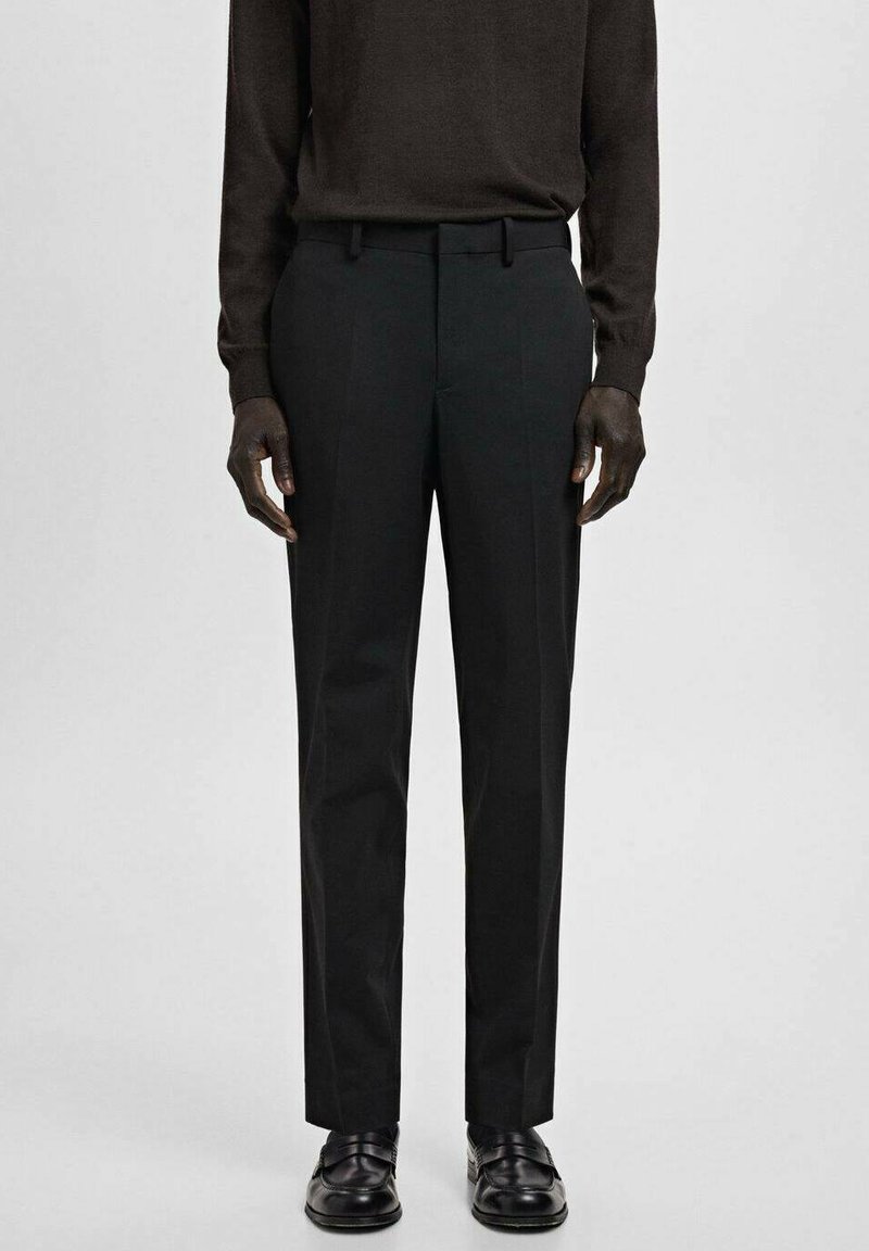 Mężczyzna ubrany w czarne spodnie garniturowe, czarne mokasyny i ciemnobrązową koszulę z długim rękawem, stojący na tle jednolitego jasnoszarego tła.