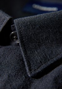 Collare in tessuto blu scuro con texture visibile, dettaglio dei bottoni e un taglio pulito. Le cuciture sono ordinate, a testimonianza di un'alta qualità artigianale.
