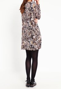 Sukienka z długim rękawem z wirującym wzorem paisley w brązowym, kremowym i czarnym kolorze. Wykonana z miękkiego materiału, zestawiona z czarnymi rajstopami i butami.