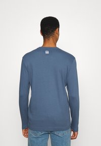 Pitkähihaisen sinisen puuvillapaidan, jossa on pyöreä kaula-aukko, takana on etiketti. Paita on yhdistetty vaaleansinisiin farkkuihin.