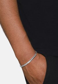 Bracciale d'argento con pietre circolari trasparenti, incastonate in fila, indossato su un polso. Lo sfondo presenta una camicia nera e pantaloni.