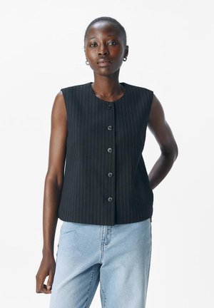 Femme portant un haut noir à rayures verticales sans manches et un jean bleu clair, posant devant un fond blanc.