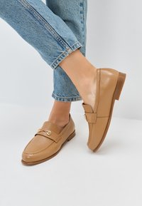 Mocassini in pelle color tan con punta arrotondata e tacco basso, dotati di un accento in metallo dorato sulla fascia. Indossati con jeans azzurri chiari risvoltati.
