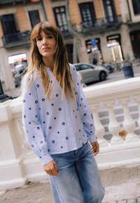 Giovane donna con capelli lunghi, indossa una camicia azzurra a fantasia con bottoni e jeans, in piedi su un marciapiede cittadino vicino a una balaustra bianca.