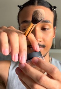 Bocal cosmétique en verre transparent avec un couvercle noir, tenu par une main. Un pinceau à maquillage avec un manche doré et des poils doux est positionné au-dessus.