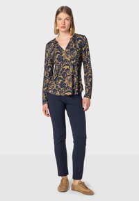 Langärmliges navy-blaues Oberteil mit goldenem Blumenmuster, V-Ausschnitt, kombiniert mit navy-blauen Hosen und braunen Schuhen, vor einem schlichten Hintergrund stehend.