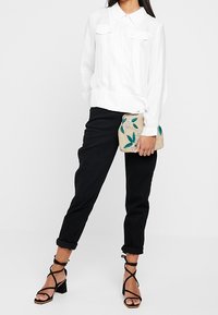 Kvinde iført en hvid bluse med lange ærmer, sorte op-rullede bukser, sorte hæle med stropper, der holder en beige clutch med grønne blade broderet på.