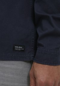 Giacca blu navy realizzata in tessuto morbido, con un'etichetta con testo nero. Il design ha una vestibilità comoda e polsini standard.