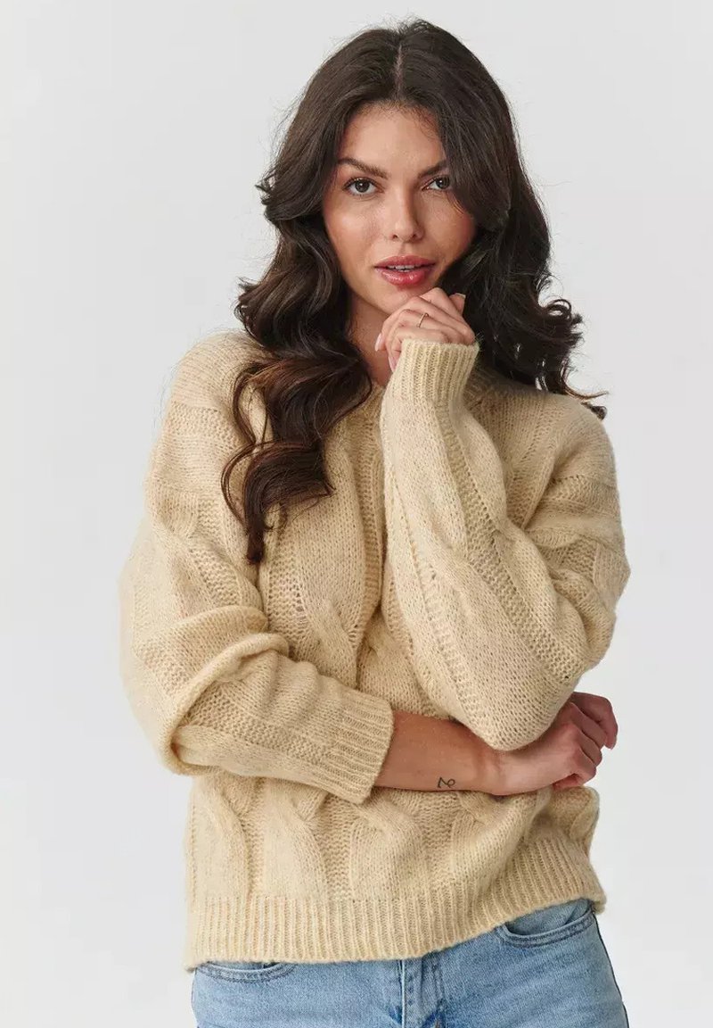 Beżowy sweter z dzianiny w warkocze o luźnym kroju, ściągacze i dół wykończone prążkowo, miękka faktura, noszony na jasnoniebieskie dżinsy.