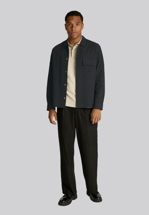 Camicia scura con bottoni e una tasca sul petto, indossata sopra una polo chiara, abbinata a pantaloni e scarpe nere. Tessuto opaco e vestibilità casual.