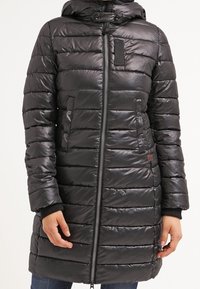 Černý prošívaný dlouhý péřový kabát s kapucí, předním zipem, zapínacími kapsami na flopu a žebrovanými manžetami, který nosí stojící osoba.