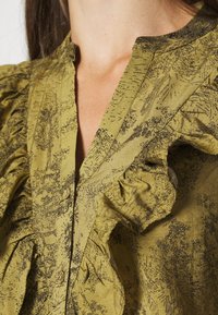 Blusa verde oliva con dettagli arricciati, caratterizzata da un scollo a V e chiusura con bottoni. Il design include stampe botaniche nere e tessuto testurizzato.