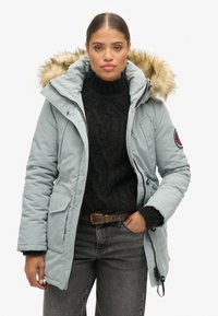 Parka isolante blu chiaro con cappuccio bordato in pelliccia, chiusura con zip e due tasche frontali, abbinata a un maglione nero a maglia cable e jeans scuri.