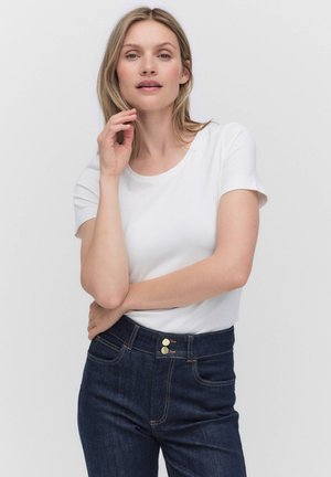 Blonde Frau trägt ein weißes Kurzarmshirt und dunkelblaue, hochtaillierte Jeans, posiert mit einer Hand nahe ihrem Gesicht.