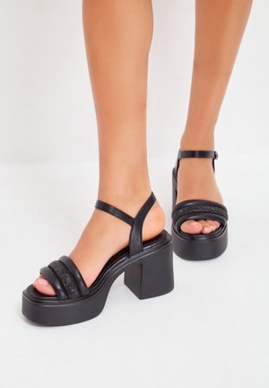 Sandales plates noires à plateforme avec une finition brillante, présentant deux sangles noires texturisées et une sangle autour de la cheville. Design de talon carré.