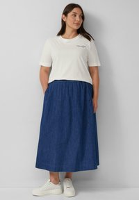 Weißes Baumwoll-T-Shirt mit kurzen Ärmeln und einem kleinen Druckdetail, kombiniert mit einem langen, dunkelblauen Jeansrock und weißen Sneakers.